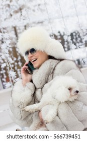 Beautiful Woman Making A Phone Call While Holding Her Dog Outdoors. Winter Time. Woman Wearing A White Fur Coat. Christmas Decorations In The Background.