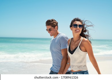 Beautiful woman with man wearing sunglasses walking on beach.. Young couple enjoying honeymoon after marriage at sea. Happy casual couple holding hands and walking at the beach with copy space. - Powered by Shutterstock