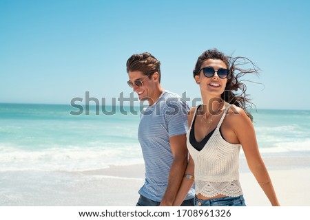 Beautiful woman with man wearing sunglasses walking on beach.. Young couple enjoying honeymoon after marriage at sea. Happy casual couple holding hands and walking at the beach with copy space.