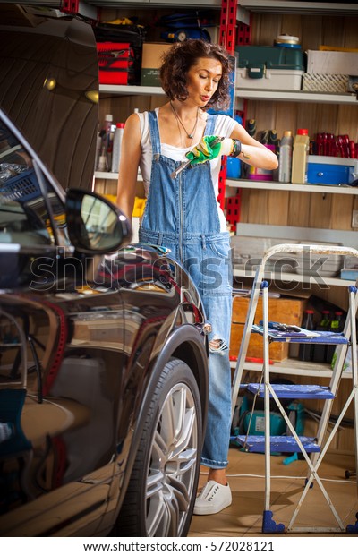beautiful woman\
mechanic in blue overalls looks at wristwatch. Car body shop in the\
garage and a female car\
mechanic