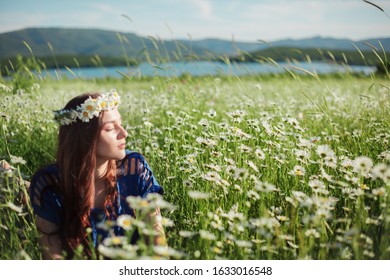 Beautiful woman with nude makeup posing in the nature flowers field at the bacground beatiful views with mountains. Female holding bouquet with wild growing daisies and wreath.