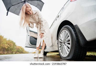 Beautiful Woman On A Road Near Her White Car With Punctured Car Tire. Female Driver Looking To Deflated Tyre, Holding Umbrella.