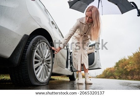 Beautiful woman on a road stopped her white car with punctured car tire. Female driver looking to deflated tyre, holding umbrella.