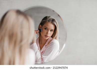 Beautiful Woman In Pajamas In Front Of Mirror, Holding Brush And Applying Makeup, Face Powder Or Bronzer In Bathroom, Free Space. Cosmetics For Makeup Concept