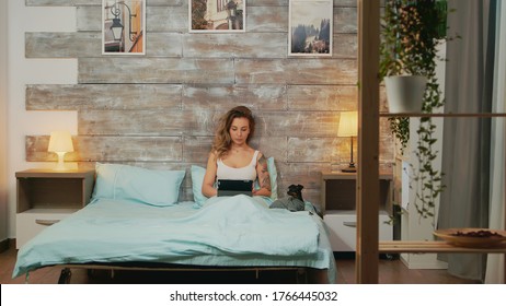 Beautiful woman in pajamas using tablet computer in bed. Little dog beside her