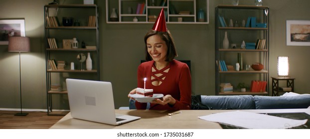 Beautiful Woman In A Party Cap Lights A Candle On A Cake, Having An Online Birthday Party With Friends. Stay Home, Quarantine Life. Shot With Anamorphic Lens