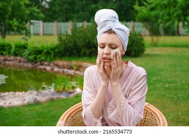Beautiful woman with perfect skin doing makeup and cleansing skin on her face with cotton pillow after shower in towel on her head while sitting in chair garden at home in fresh air Skin care concept
