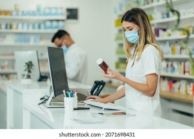 Beautiful Woman Pharmacist With Protective Mask On Her Face Holding Drugs And Using Computer While Working At A Pharmacy