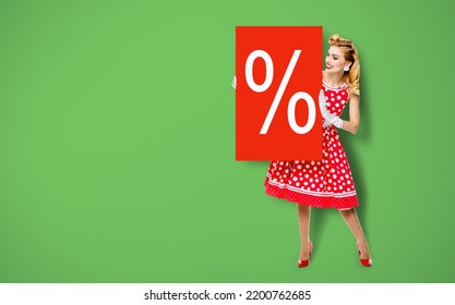 Beautiful Woman In Pinup Dress Holding, Showing Red Board With Procents % Sign. Full Body Pin Up Girl With Advertising Offer, Isolated Over Green Background. Sales, Discounts Rebates Ad Concept.