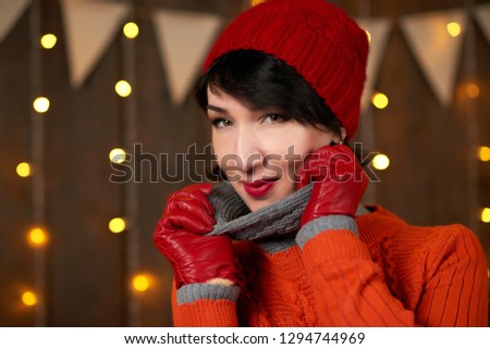 Beautiful woman is posing on dark wooden background, christmas lights and flags, holiday concept. Dressed in red knitted hat and sweater.