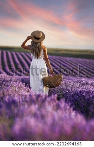 Beautiful woman at purple lavender field with violet blossom lavanda flowers. French young blond girl in provence lavender farm wears white dress and hat with wicker basket at sunset meadow. 