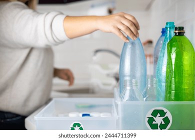 A beautiful woman puts a plastic bottle into a box with a recycling sign. Sorting plastic at home in the kitchen. A woman puts plastic bottles in a box for recycling. - Powered by Shutterstock