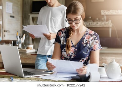 Beautiful woman reads documents attentively, prepares financial report, calculates figures and works on generic portable laptop computer. Family couple mange budget, plan to buy something expensive