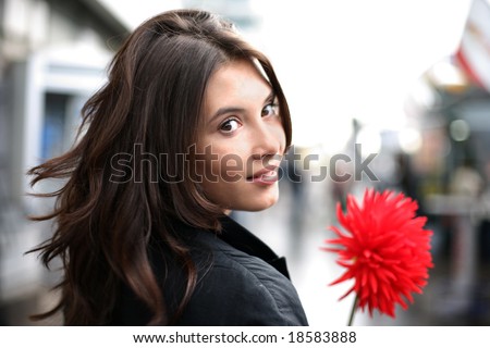 Beautiful woman with red flower walking down street, looking back. Shallow DOF.