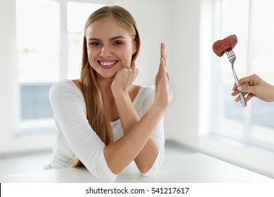 Beautiful Woman Refuses To Eat Red Meat. Female Hand Holding Fork With Piece Of Beef Steak, Proposing It To Girl. Attractive Female With Stop Hand Sign. Vegetarian Nutrition Concept. High Resolution