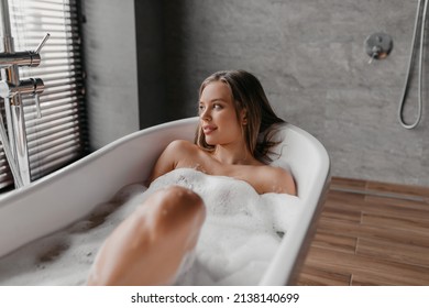 Beautiful Woman Relaxing In Bathtub Filled With Foam, Relaxed Lady Sitting In Hot Bath And Looking At Window, Resting In Modern Bathroom Interior. Domestic Spa Concept