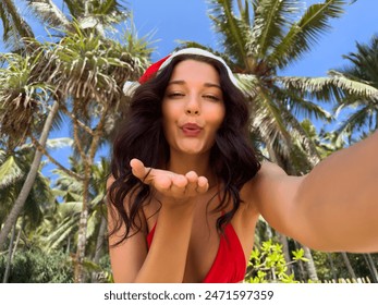 Beautiful woman in a Santa hat blowing a kiss towards the camera on a tropical beach. The vibrant palm trees and clear blue sky create a festive and joyful atmosphere, perfect for holiday-themed - Powered by Shutterstock
