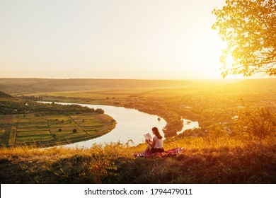 A beautiful  woman sits on a hill and reads a book against the background of a panorama of mountains, river and amazing sunset. Relaxing moment.