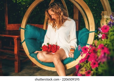 beautiful woman sits on a wooden swing with a bouquet of red roses in her hand. - Shutterstock ID 2206509195