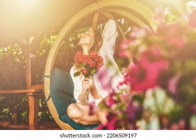 beautiful woman sits on a wooden swing with a bouquet of red roses in her hand. - Shutterstock ID 2206509197