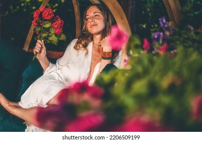 beautiful woman sits on a wooden swing with a bouquet of red roses in her hand. - Shutterstock ID 2206509199