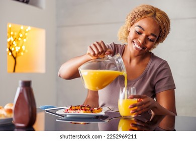 Beautiful woman sitting at dining room table pouring glass of freshly squeezed orange juice while having breakfast at home in the morning - Powered by Shutterstock