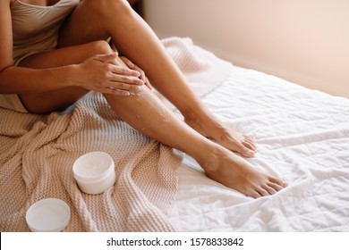 Beautiful woman sitting on bed and applying cream on legs. Young woman applying body lotion on legs