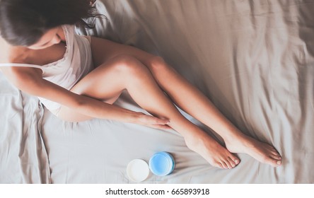 Beautiful woman sitting on bed and applying cream on legs