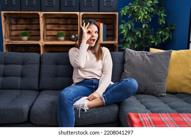 Beautiful Woman Sitting On The Sofa At Home Smiling Happy Doing Ok Sign With Hand On Eye Looking Through Fingers 