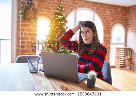 Beautiful woman sitting at the table working with laptop at home around christmas tree confuse and wonder about question. Uncertain with doubt, thinking with hand on head. Pensive concept.