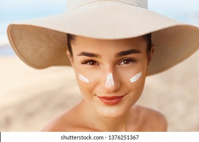 Beautiful Woman smile applying sun cream on face. Skin and  Body  care. Sun protection. Girl in bikini holding sunscreen bottle on the beach. Female in hat smear moisturizing lotion on skin
