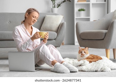 Красивая женщина в мягких тапочках с чашкой кофе и милый кот сидит на ковре дома