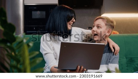 Beautiful woman with son using laptop at home. Happy mom and preteen school child sitting on sofa at home kitchen, watching cartoons online or play video game. Joyful family