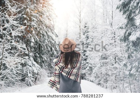 Beautiful woman standing among snowy trees in winter forest and enjoying first snow. Wearing hat, plaid scarf and coat. Wanderlust and boho style