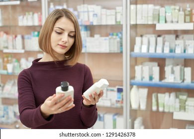 Beautiful woman standing in drugstore and choosing between two goods. Pharmacy customer holding in hands two white cosmetic bottles. Consuming of medical products.