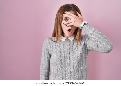 Beautiful woman standing over pink background peeking in shock covering face and eyes with hand, looking through fingers with embarrassed expression. 