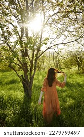 beautiful, a woman stands in a long orange dress, in the countryside, near a flowering tree, during sunset, stands with her back to the camera, holding her hands loosely near her head - Shutterstock ID 2279799497
