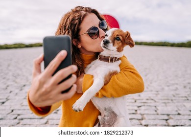 A beautiful woman staying in a park with her cute little dog. She is taking a self-portrait photo with the phone while kissing her pet. Technology lifestyle with pets