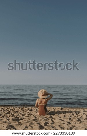 Beautiful woman in straw hat and swimsuit sitting on sand at beach and watching at sea and sky. Aesthetic summer fashion concept
