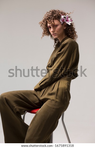 Beautiful woman in a stylish green\
suit is sitting on a chair. Curly hair. Hairstyle with orchids.\
Fashion, style and beauty. One man on an isolated\
background.