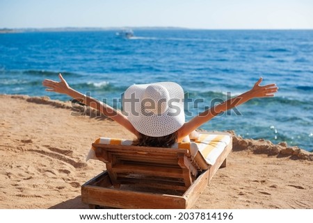 Beautiful woman sunbathing on a beach at tropical travel resort, enjoying summer holidays raising her hands up. Young woman lying on sun lounger near the sea. Happy serene woman