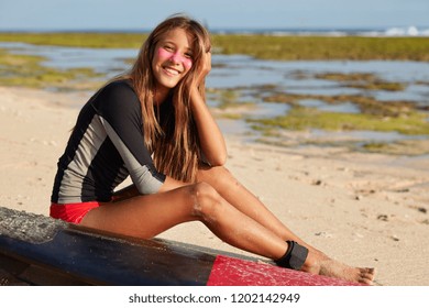 Beautiful woman surfer dressed in bathingsuit, has protective surf zinc on face, shows slender legs, has legrope or cord attached to tail of surfborad, thinks about safety during hiting waves - Powered by Shutterstock