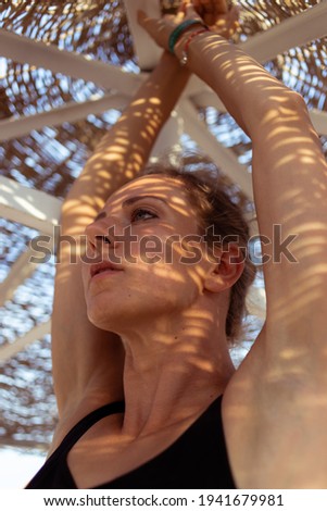 Beautiful woman in swimsuit standing under a beach umbrella with contrasting shadows from branches on the face. Summer vacation concept