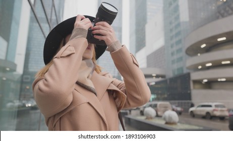 Beautiful woman takes urban winter landscape. Action. Stylish woman shoots urban landscapes on professional camera. Woman photographer takes modern urban high-rise buildings in snow
