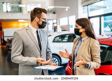 Beautiful  Woman Talking With Salesman At Car Showroom About New Car She Wants To Buy. They Are Wearing Face Protective Masks Due To Covid-19 Pandemic.