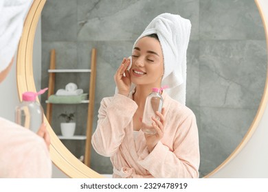 Beautiful woman in terry towel removing makeup with cotton pad near mirror indoors - Powered by Shutterstock