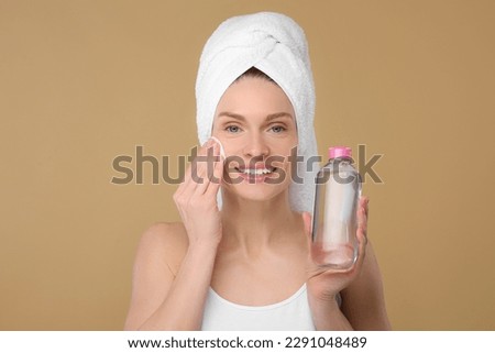 Beautiful woman in terry towel removing makeup with cotton pad on beige background