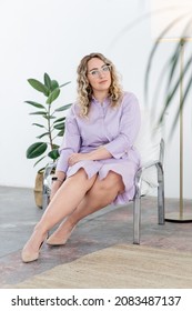 A Beautiful Woman Therapist With Glasses Is Sitting In A Chair And Looking At The Camera. On The Background Of A Potted Plant. There Is A Tall Floor Lamp Nearby. Look Into The Camera. Plus Size Model