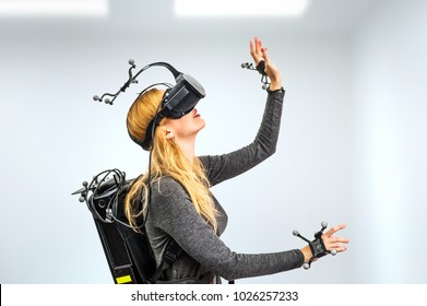 Beautiful Woman Touching Air During VR Experience In Room. She Wearing Virtual Reality Glasses Or Goggles, Processor In Backpack On Back And Sensors For Tracking On Head And Hands