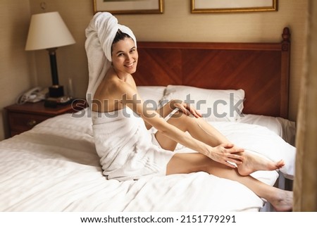 Beautiful woman in towel on head and towel on breast sitting on bed in hotel room and applying cream on legs. Young woman applying body lotion on legs. 